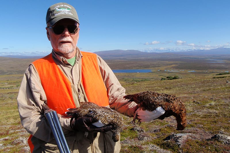 A hunter in an orange vest poses with two captured ptarmigans in front of an expansive field.