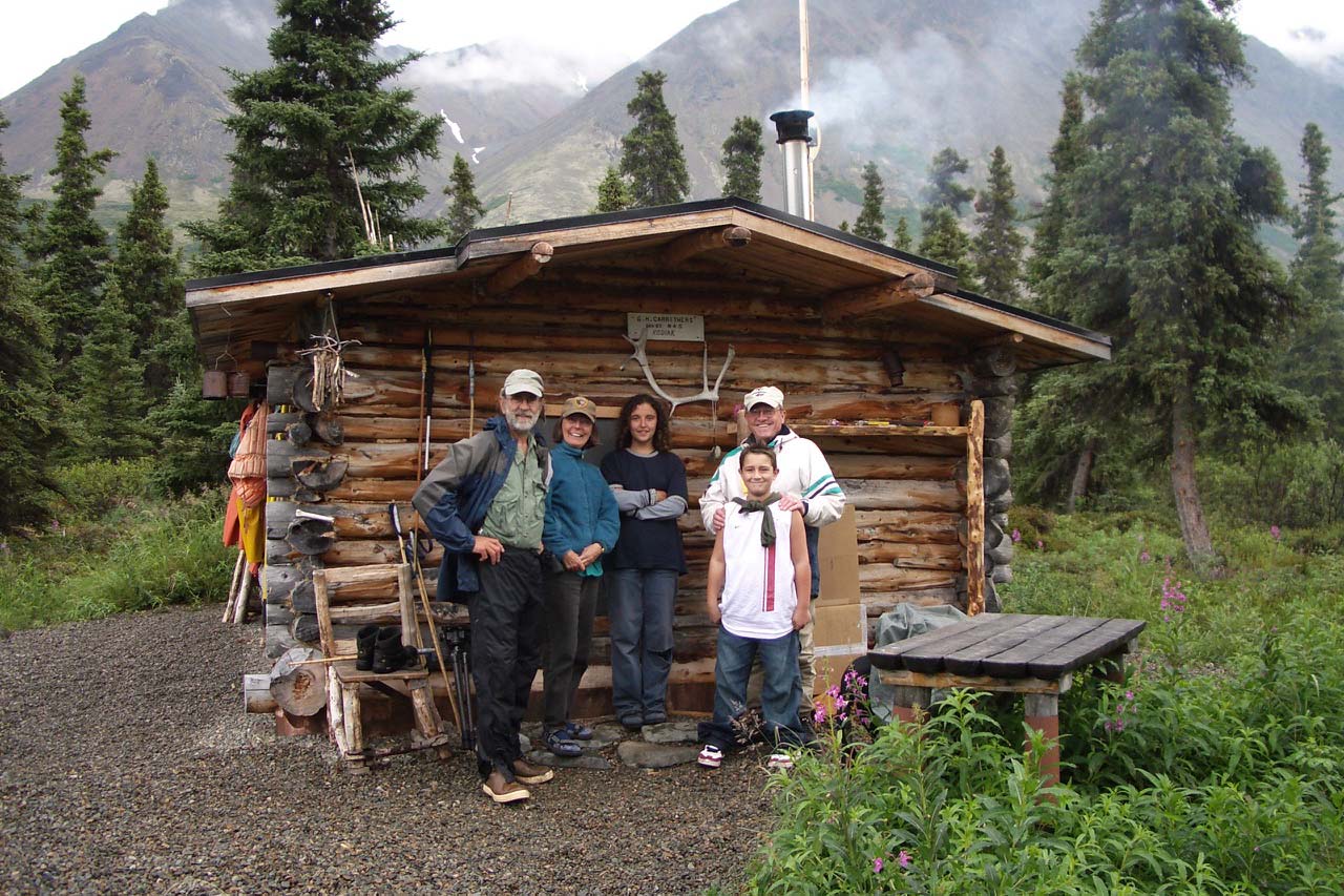 A group of people stand in front of a quaint log cabin beneath high mountains.